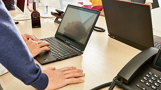 Schreibtisch mit verschiedenen Computern und Menschen, die diese bedienen und sich dabei austauschen.